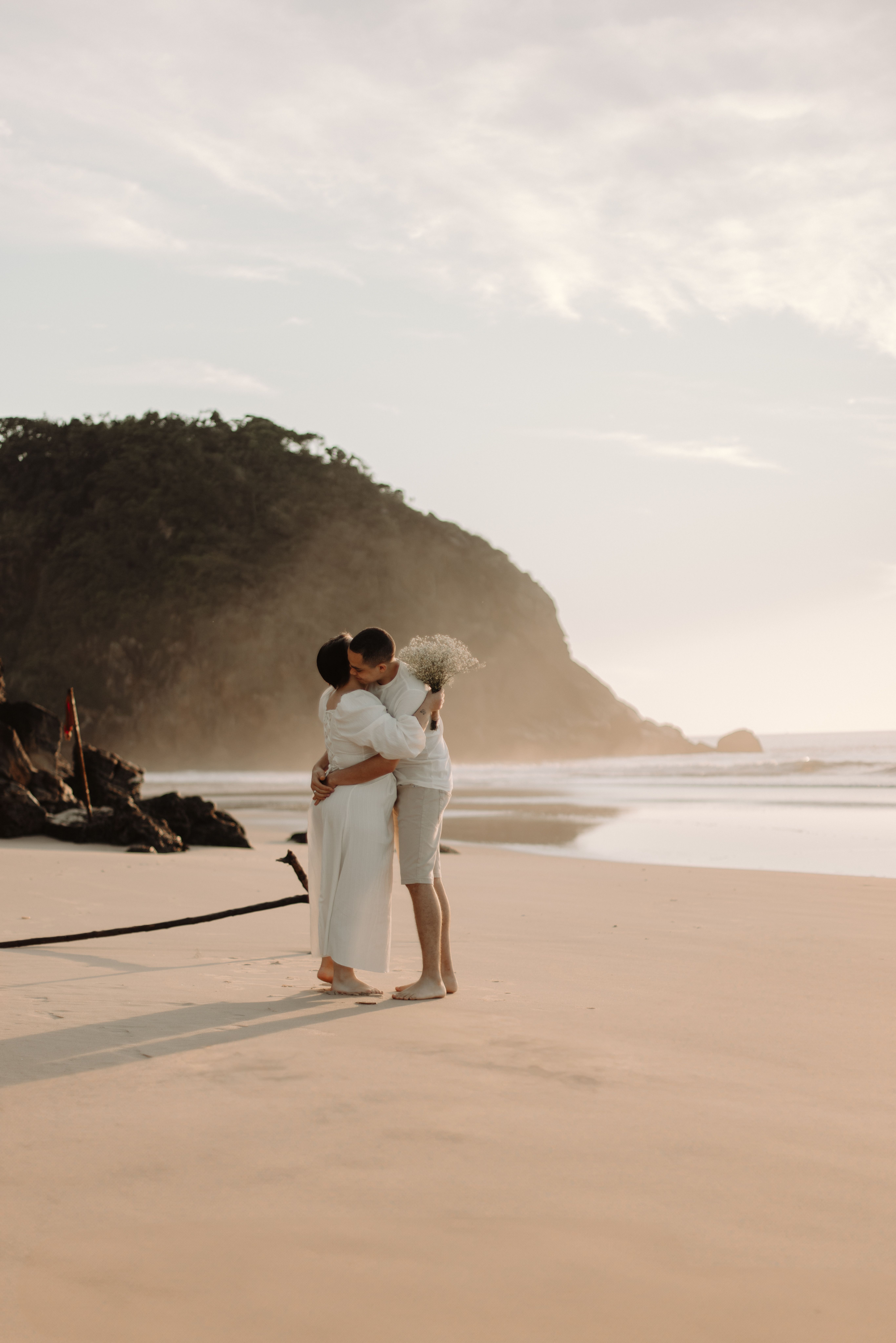 Couple embracing barefoot on a serene beach at sunrise, candid intimate elopement moment, warm coastal light
